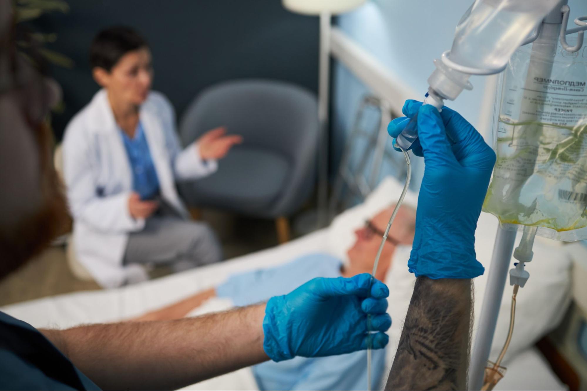 A man is lying down on a bed while a nurse is prepping an IV.