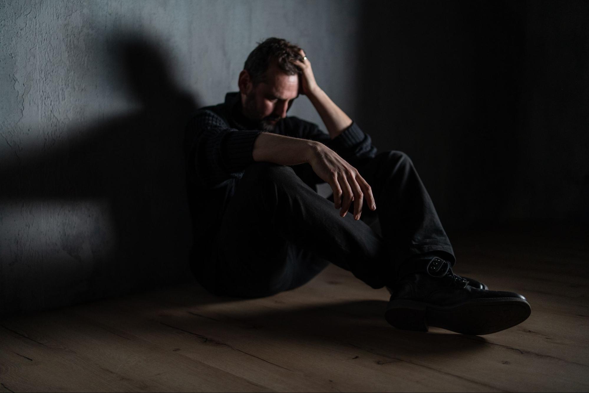 A man with low testosterone feels depressed and sits on the floor of a dark room.