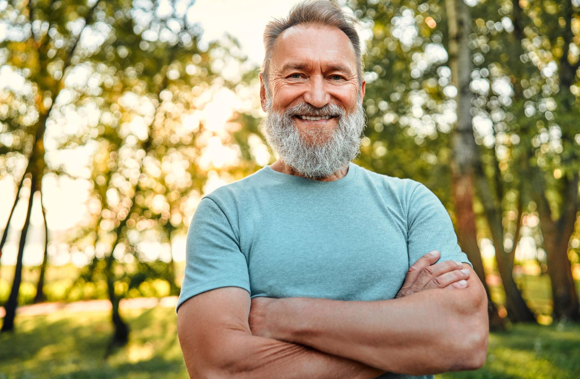 A bearded, muscular mature man stands in the forest smiling and cross-armed.