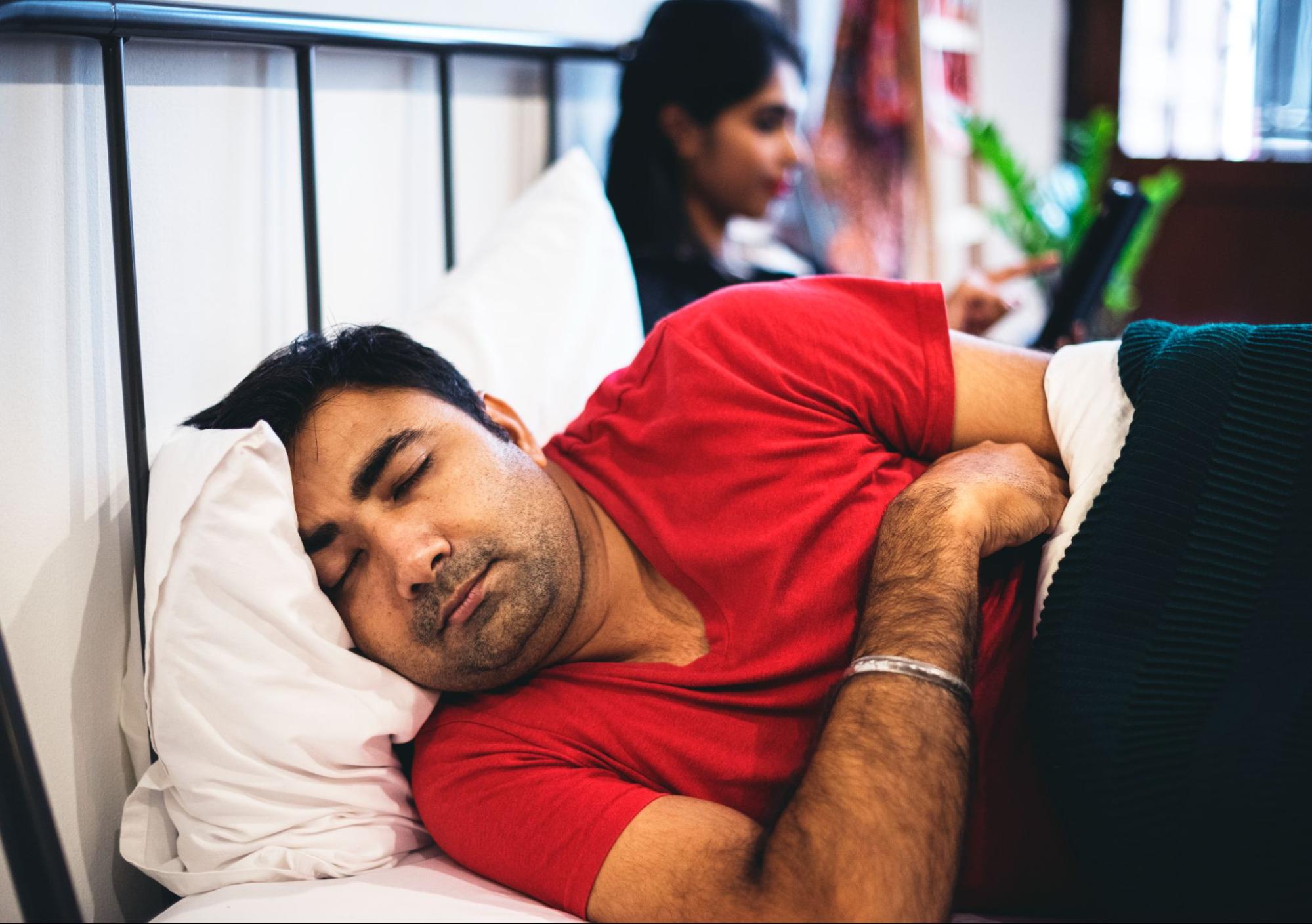 A man lies in bed with his back to his upset wife, pretending to sleep to avoid intimacy